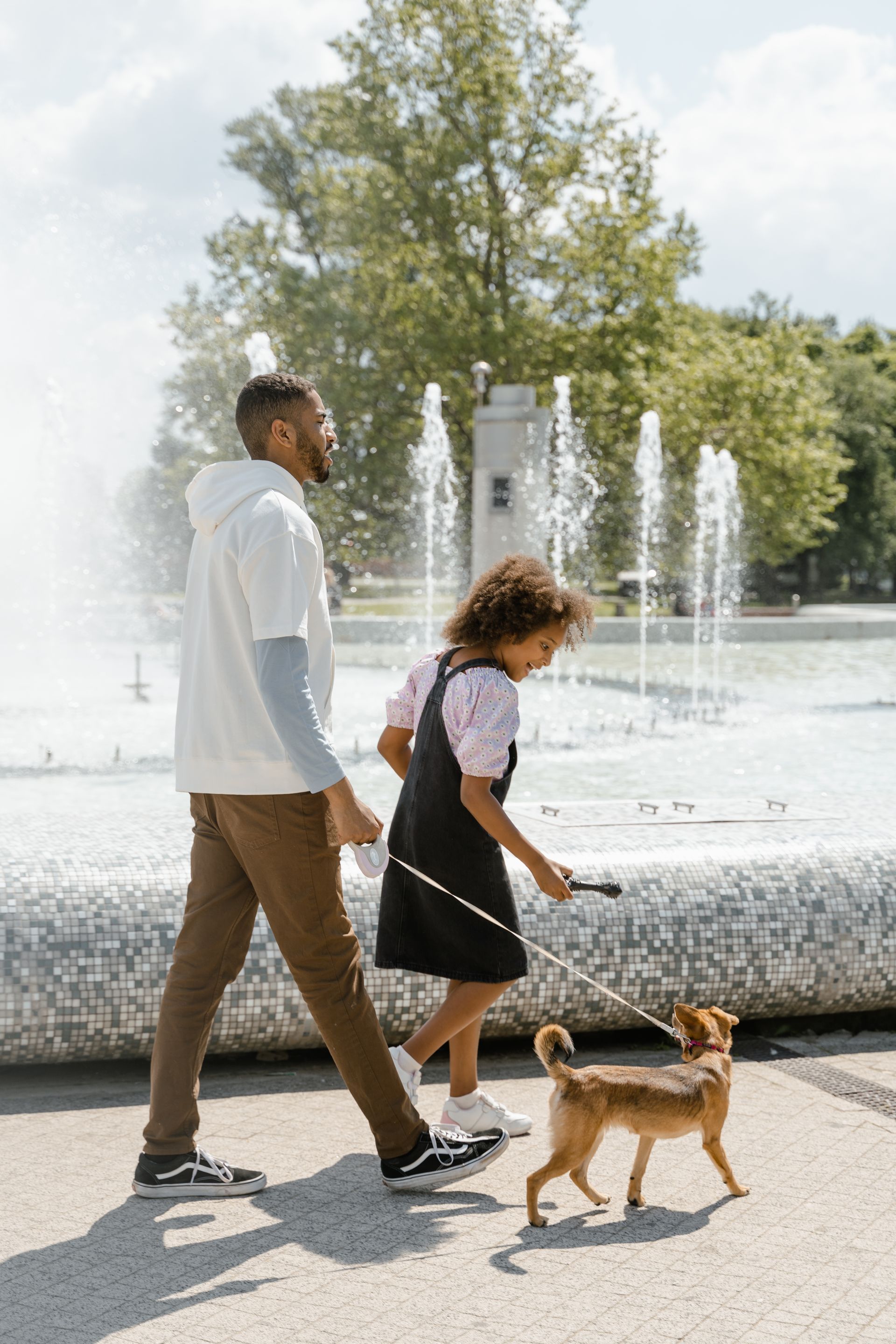 Padre e hija paseando a su perro junto a una fuente pública